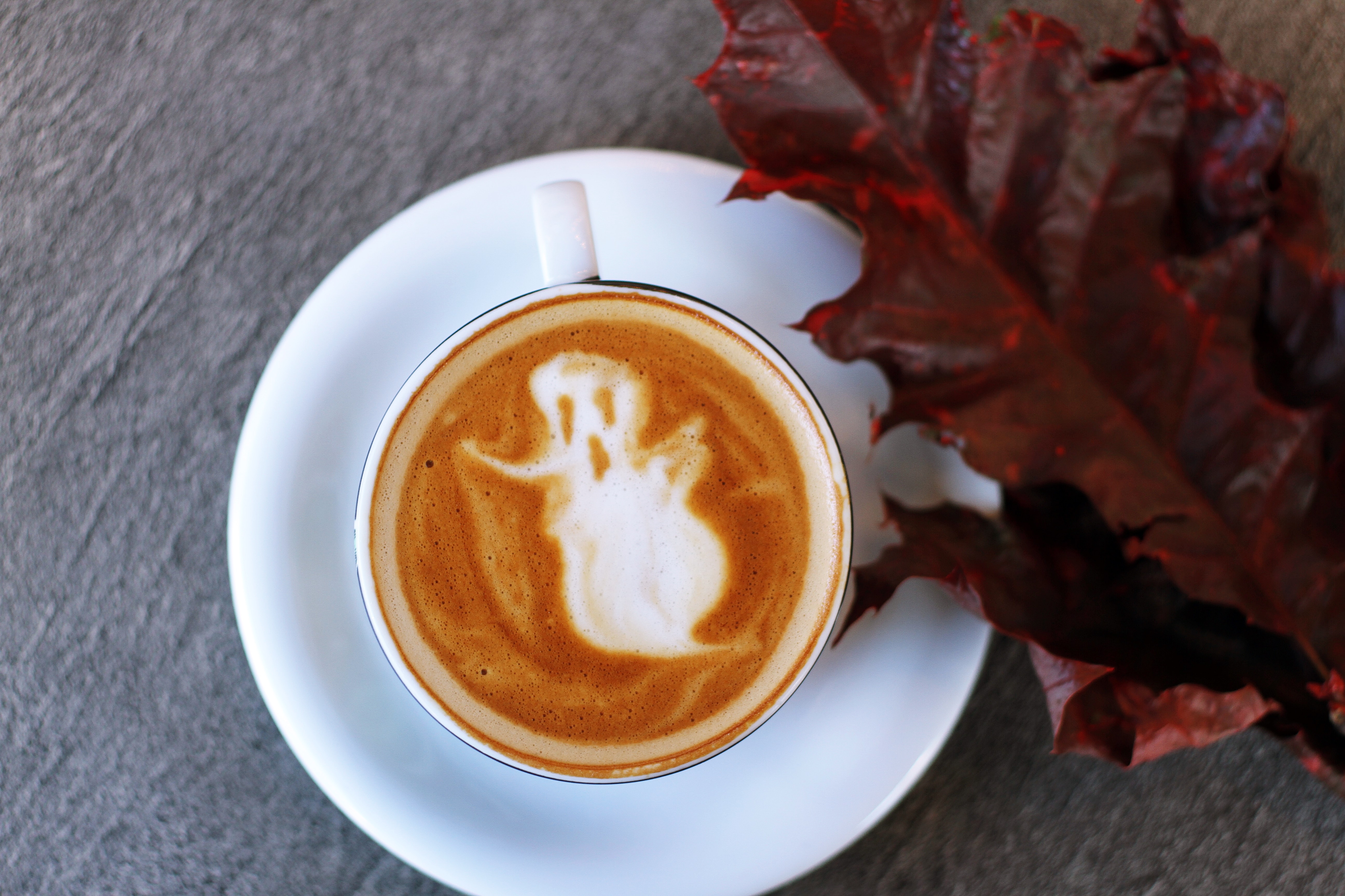 A latte with a ghost in the foam and a fall leaf on the table next to it.